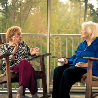 Two elderly women engaged in conversation while sitting on a porch.