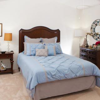 A cozy bedroom featuring a neatly made bed with blue bedding, a wooden headboard, and surrounding furniture including a nightstand with a lamp and a dresser with a decorative mirror.