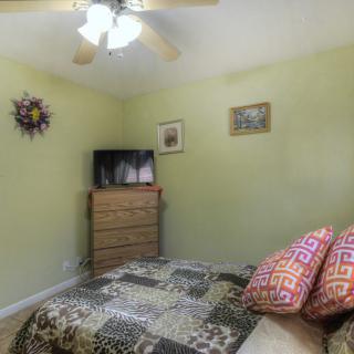 A cozy bedroom interior featuring a bed with patterned bedding, a wooden dresser topped with a television, and a ceiling fan overhead.