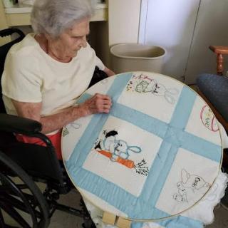 An elderly woman in a wheelchair is focused on her embroidery work at a table.
