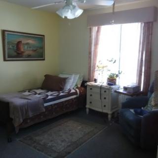 A cozy bedroom featuring a single bed, an armchair, a dresser by the window with curtains, and a painting on the wall.
