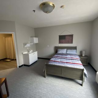 A minimalist bedroom featuring a neatly made bed, a side table with a lamp, and a window providing natural light.