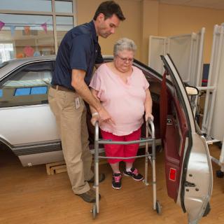 A caregiver assists an elderly woman using a walker to exit a car in a healthcare setting.