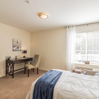A cozy bedroom with a neatly arranged desk and large window letting in natural light.