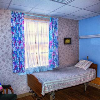 A simple hospital room with a single bed, floral curtains, and a wooden chair.