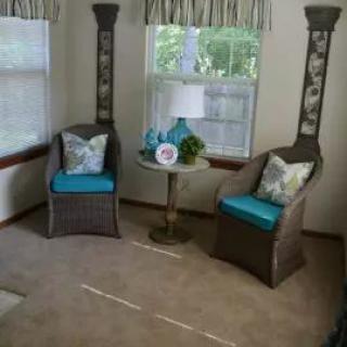 A cozy sitting area with two wicker chairs and a small round table between them, placed near windows with patterned curtains.