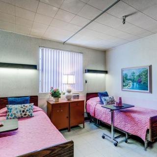 A hospital room featuring twin beds with pink bedding, bedside tables, and a decorative painting on the wall.