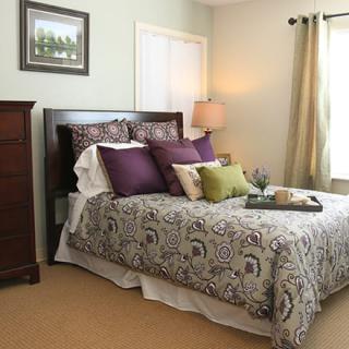 A cozy bedroom featuring a decorated bed with patterned bedding, a lamp on the side, and natural light coming through the window.