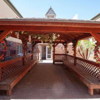 A wooden gazebo with bench seating, featuring lattice designs, located between brick buildings.