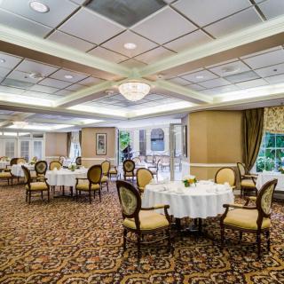 A spacious and elegant banquet hall set with round tables and upholstered chairs, featuring chandeliers and large windows.