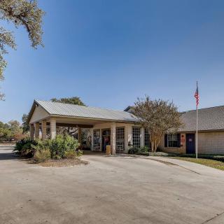 A single-story building with a covered entrance and a parking lot, surrounded by trees and featuring a flagpole.
