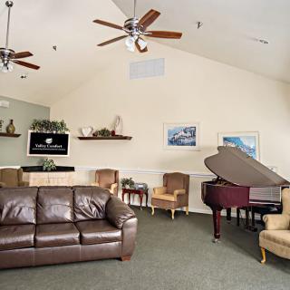 A spacious living room featuring a leather sofa, armchairs, a grand piano, and a wall-mounted television with ceiling fans overhead.