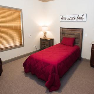 A cozy bedroom with a single bed featuring red bedding, a nightstand with a lamp, and window blinds.