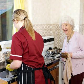 A caregiver assists an elderly woman in the kitchen, engaging in household chores together.