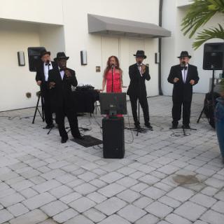 A lively outdoor performance featuring a band dressed in black suits and hats, accompanied by a female singer in a vibrant pink outfit.