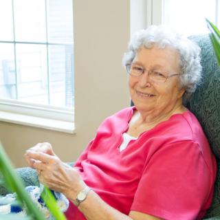 An elderly woman sitting comfortably in a chair, smiling while knitting by the window.