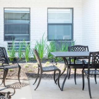 A modern patio with a set of outdoor furniture, featuring a table and chairs, surrounded by plants and large windows.
