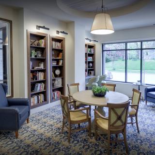 A cozy library reading area with bookshelves, a round table and chairs, illuminated by natural light from large windows.