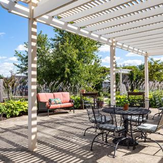 A sunny patio with a pergola overhead, featuring outdoor seating and surrounded by lush greenery.