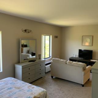 A cozy living space with a bed, dresser, sofa, and television, featuring neutral tones and natural light.