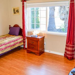 A cozy bedroom with a single bed, wooden floors, and a large window with red curtains.