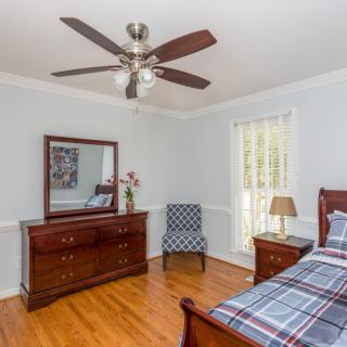 A neatly arranged bedroom with wooden furniture, featuring a bed, dresser with mirror, chair, and a ceiling fan.