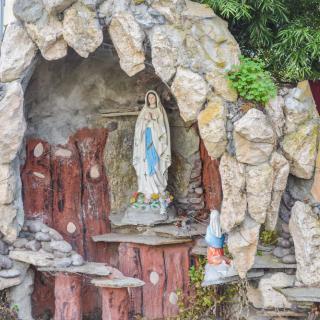 A religious grotto featuring a statue of the Virgin Mary, surrounded by stones and greenery.