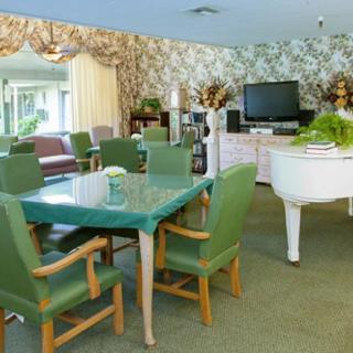 A cozy living room with a glass dining table, green chairs, a white piano, and a television set against floral wallpaper.