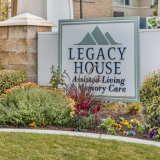 A sign for Legacy House, an assisted living and memory care facility, surrounded by landscaped greenery and flowers.