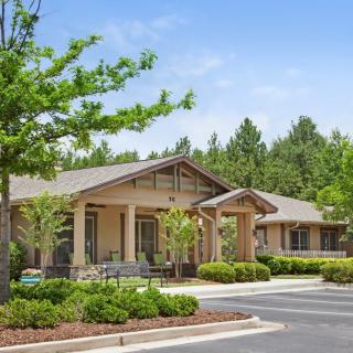A single-story building surrounded by lush greenery and trees with a parking area in front.