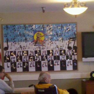 A bulletin board with a music theme is displayed indoors, with people seated in front of it.