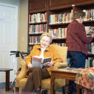 An elderly woman reading a book in a library setting, with another person browsing books.