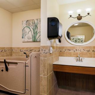 An accessible bathroom featuring a bathtub with safety handles, a sink with round mirror, and wall-mounted towel dispenser.