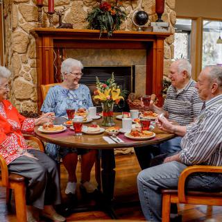A group of elderly friends enjoys a meal together around a table in a cozy room with a fireplace.