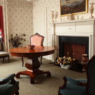 A vintage living room with antique furniture, featuring a round table, armchairs, and a fireplace.