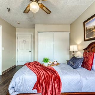 A cozy bedroom with a neatly made bed, decorative pillows, and a red throw blanket. The room features a ceiling fan, a bedside lamp, and artwork on the wall.