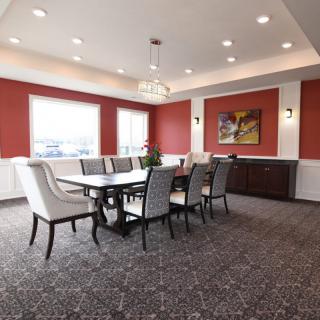 A modern dining room with a large table, surrounded by upholstered chairs, featuring red accent walls and elegant lighting.