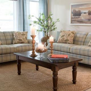 A cozy living room with plaid sofas, a wooden coffee table adorned with candles and books, complemented by a large window and wall artwork.