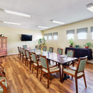 A well-lit conference room with a long table and multiple chairs, featuring a television and plants along the windows.