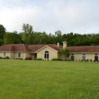 A single-story house with a red-tiled roof, surrounded by a large green lawn and trees in the background.