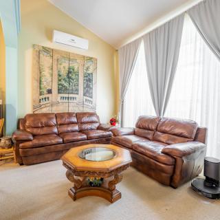 A cozy living room featuring leather sofas, a wooden coffee table, large windows with curtains, wall art, and an air conditioner.