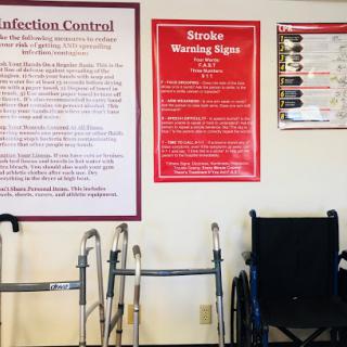 A wall in a healthcare facility displaying infection control guidelines and stroke warning signs, alongside patient mobility equipment.