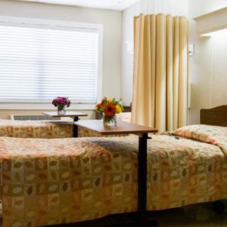 A hospital room with two beds, floral arrangements on tables, and a window with blinds.