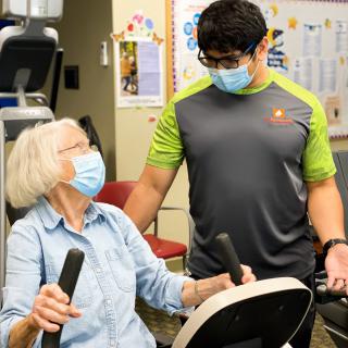A senior woman exercising on a stationary bike with the assistance of a masked trainer.