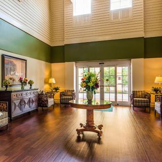 A spacious and elegantly decorated lobby with wooden floors, featuring a central table adorned with flowers, surrounded by comfortable seating and warm lighting.