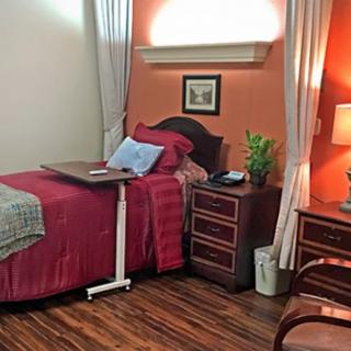 A cozy and well-furnished single hospital room with a wooden floor, bedside table, lamp, and curtains.