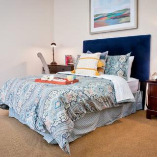A cozy bedroom featuring a bed with patterned bedding, a blue headboard, and a tidy nightstand with a lamp.