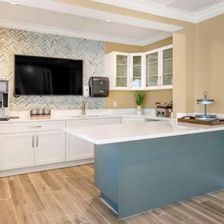 A modern kitchen featuring a central island with a white countertop, surrounded by cabinets and open shelves, equipped with a wall-mounted TV.