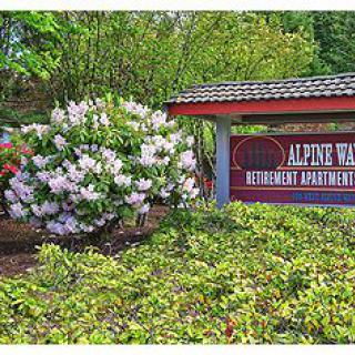 Entrance sign for Alpine West Retirement Apartments surrounded by lush greenery and blooming flowers.