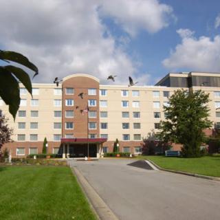 A view of a multi-story building with a driveway and surrounding greenery under a partly cloudy sky.